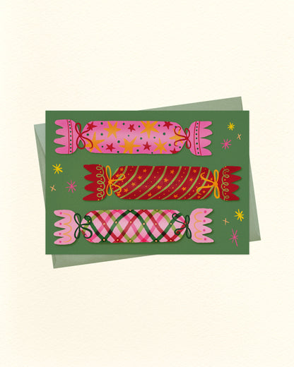 A square art print or card featuring three stylized Christmas crackers in pink, red, and green plaid patterns on a dark green background, with small yellow star accents. A light green envelope is partially visible behind the card on a warm off-white background.