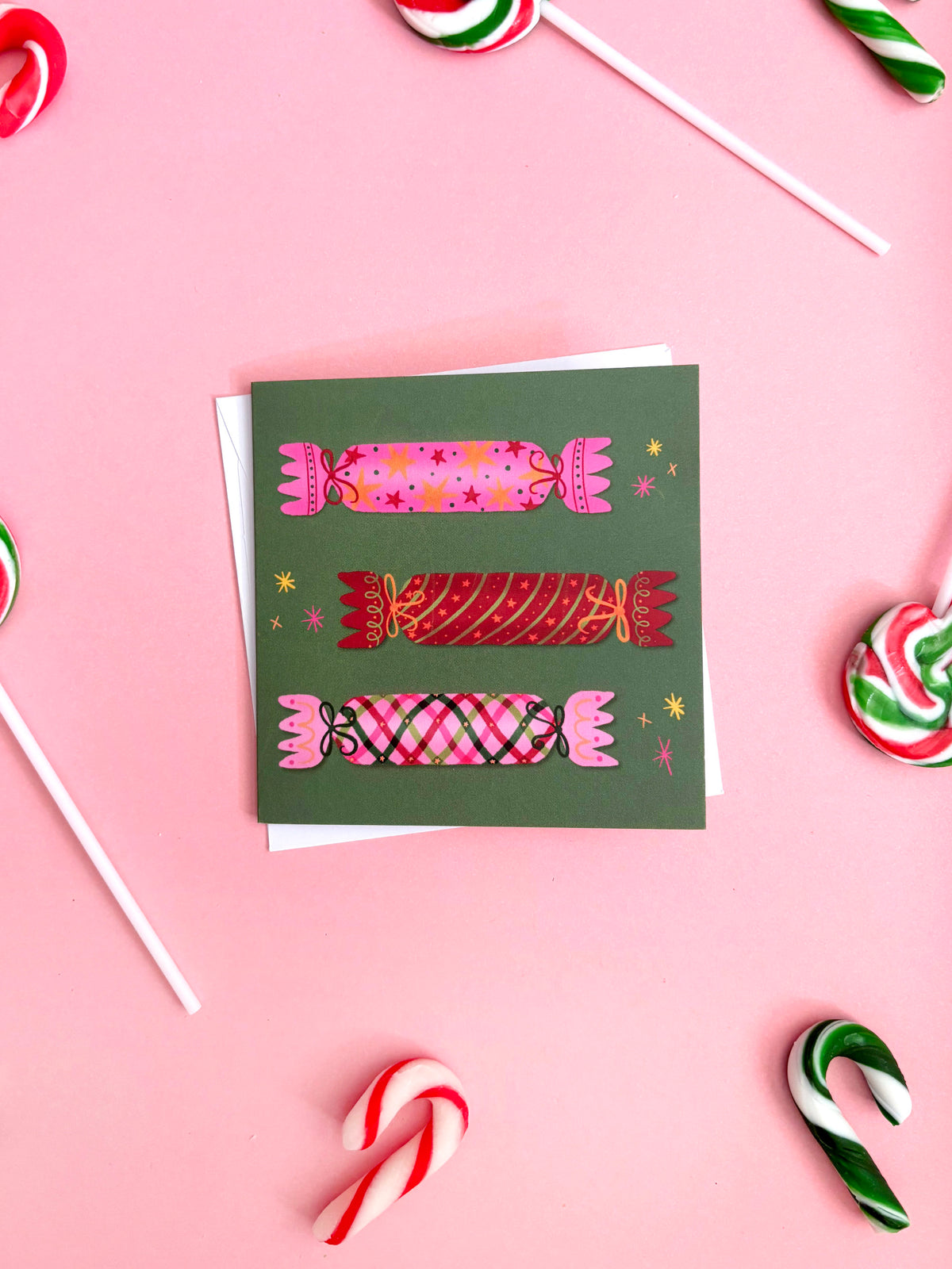A square greeting card with a dark green background and three illustrated wrapped candies in pink, red, and plaid, styled on a pink background with candy props.