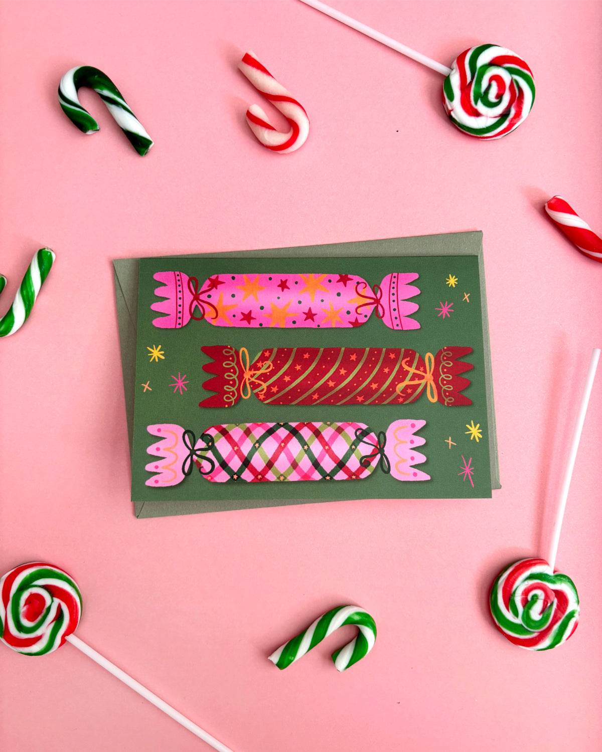 A square greeting card with a dark green background and colourful Christmas bon bons / Christmas Cracker illustrations, styled on a pink background with candy canes and lollipops around it.