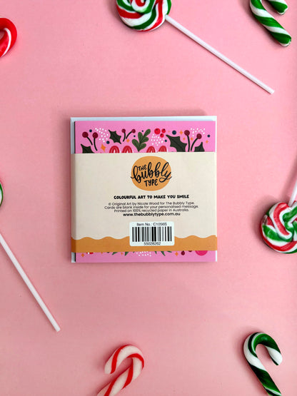 A square pack of greeting cards from The Bubble Type, featuring colourful Christmas-themed illustrations, displayed on a pink background with festive candy props.