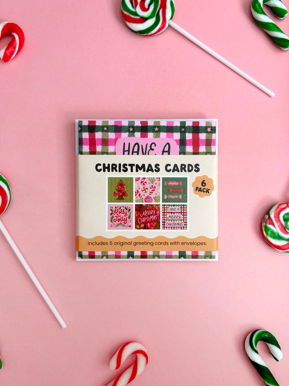 A flat-lay of a Christmas card pack on a pink background, surrounded by candy canes and lollipops. The product box is cream-coloured with a red, green, and black plaid border and displays 'HAVE A CHRISTMAS CARDS' and '6 PACK'.