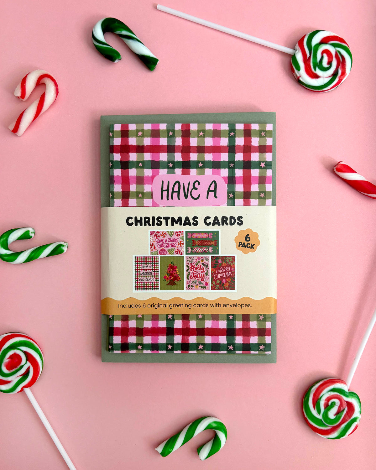 A boxed set of Christmas greeting cards arranged on a pink background with candy cane and lollipop props around it. The package front shows a red, green, and white plaid pattern with the text 'CHRISTMAS CARDS' and '6 packs'.