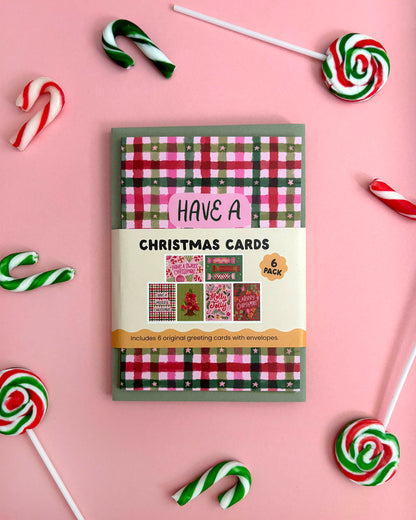 A boxed set of Christmas greeting cards arranged on a pink background with candy cane and lollipop props around it. The package front shows a red, green, and white plaid pattern with the text 'CHRISTMAS CARDS' and '6 packs'.