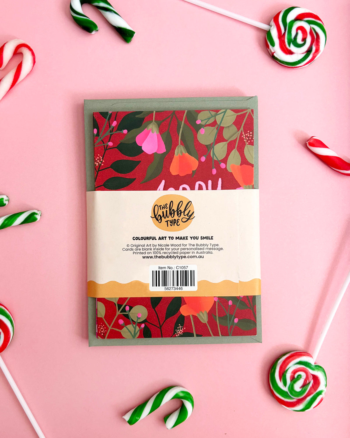 A greeting card with a colourful floral pattern, wrapped in a cream paper band labeled 'the bubbly type', arranged on a pink background with candy cane props.
