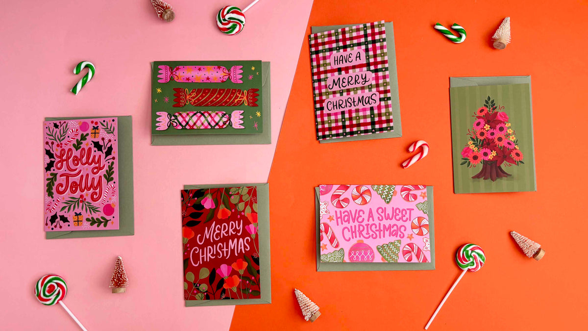 Five Christmas greeting cards arranged on a split pink and orange background with festive candy and small christmas decorations around them. The cards feature festive patterns including plaid, florals, candy motifs, and a tree illustration.
