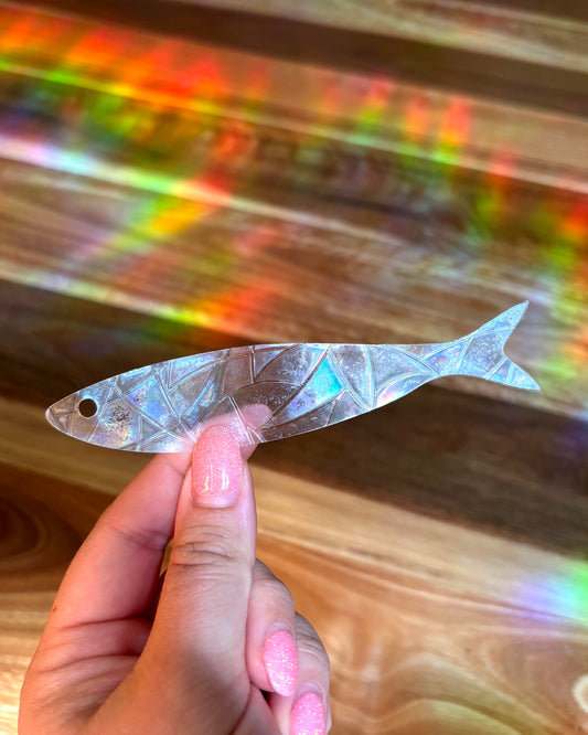 Hand holding a decorative fish-shaped sun catcher sticker with colourful rainbow light rays blurred in the background 