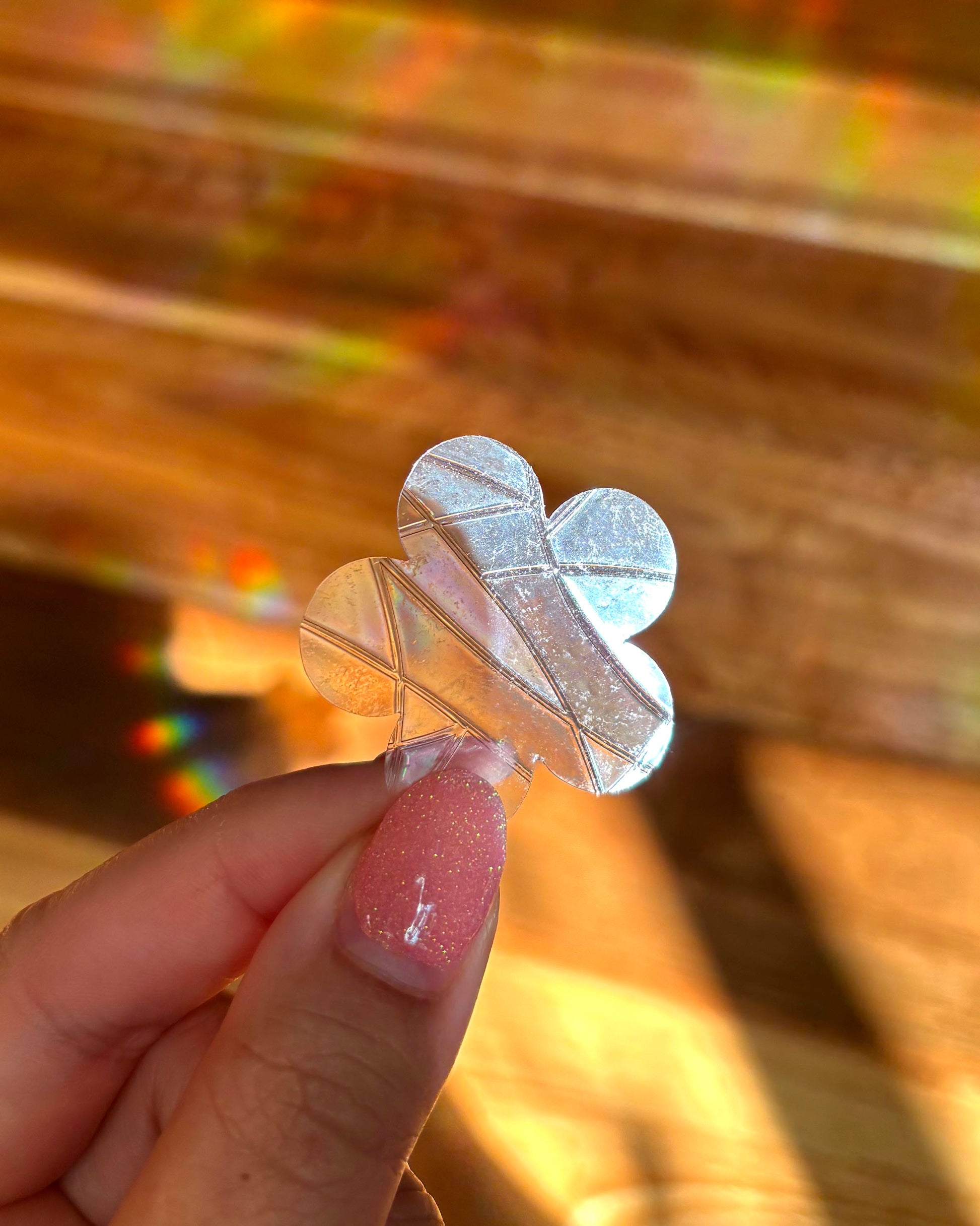 Clear flower-shaped sun catcher window decal held by a hand with pink nail polish against a blurred background with rainbow lights.