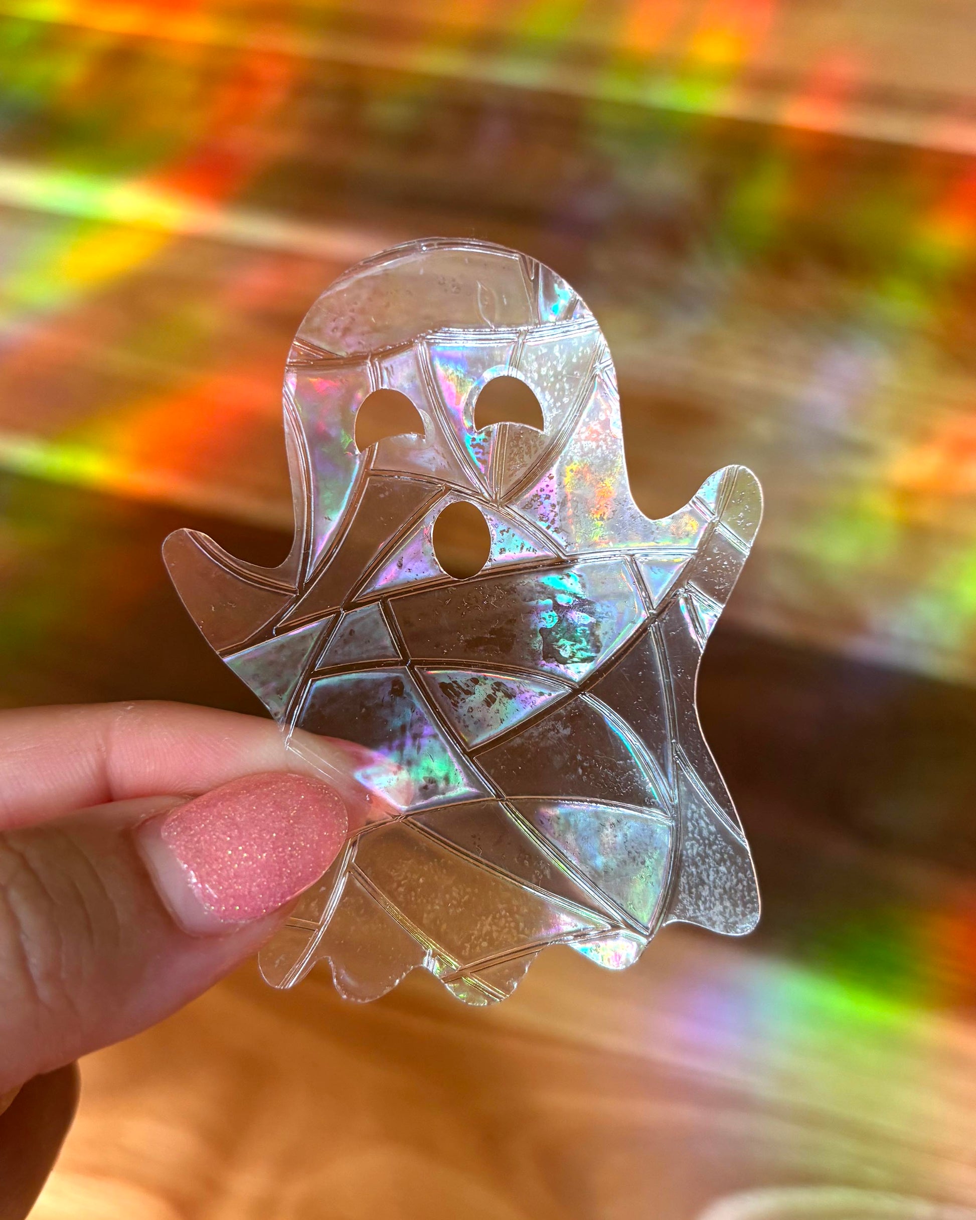 Holographic ghost-shaped sun catcher window sticker held by a hand with a multicolored background