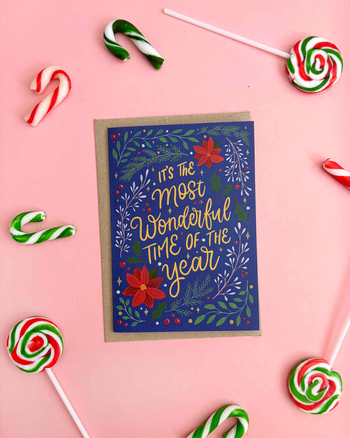 A Christmas greeting card with a navy blue background, gold script text reading 'It's the most wonderful time of the year,' and red and green botanical illustrations, placed on a pink surface with candy canes and lollipops around it.