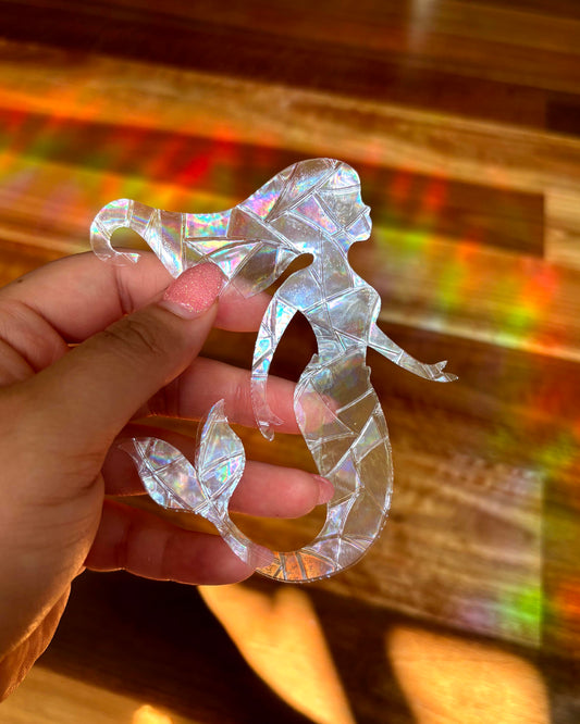 An iridescent mermaid shaped sun catcher window sicker held by a hand in front of a rainbow light filled, blurred background.