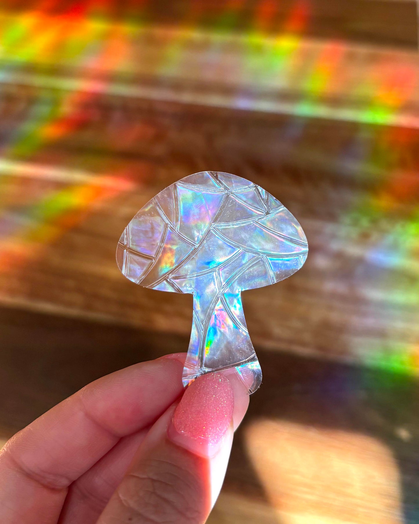 Iridescent mushroom-shaped sun catcher window cling sticker held by a hand with a colourful, rainbow lights blurred background.