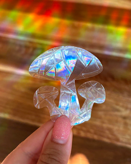 Iridescent mushroom-shaped sun catcher window sticker held by a hand with glittery nail polish against a coluorful background.