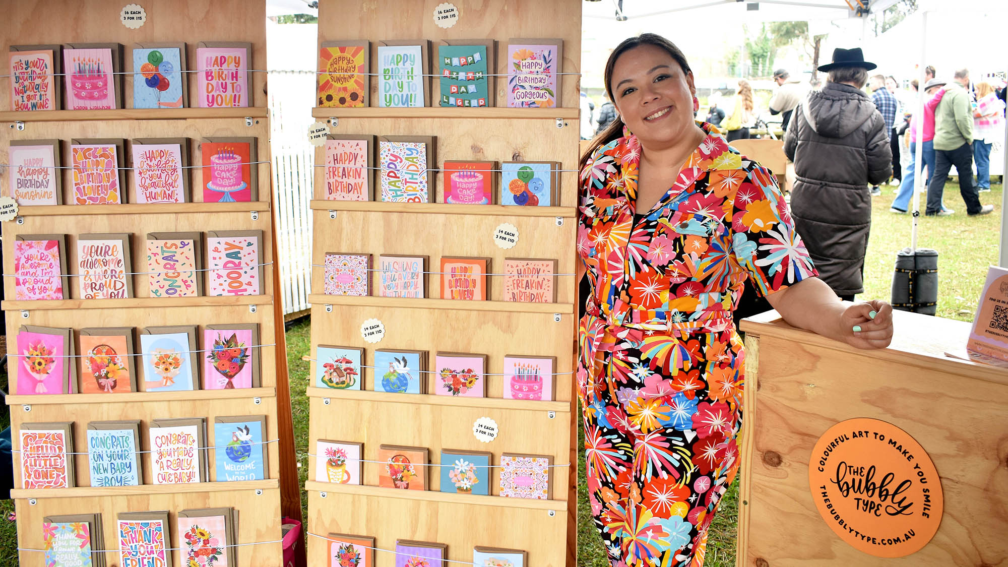 Nicole standing inside her market stall. She is wearing a colourful jumpsuit and has 2 tall wooden card stands full of her original art cards next to her.