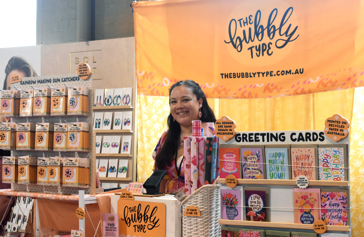 A smiling woman called Nicole stands behind a retail display booth branded 'The Bubbly Type', with greeting cards, sun catchers, and printed items arranged on shelves and tables under a large orange banner.
