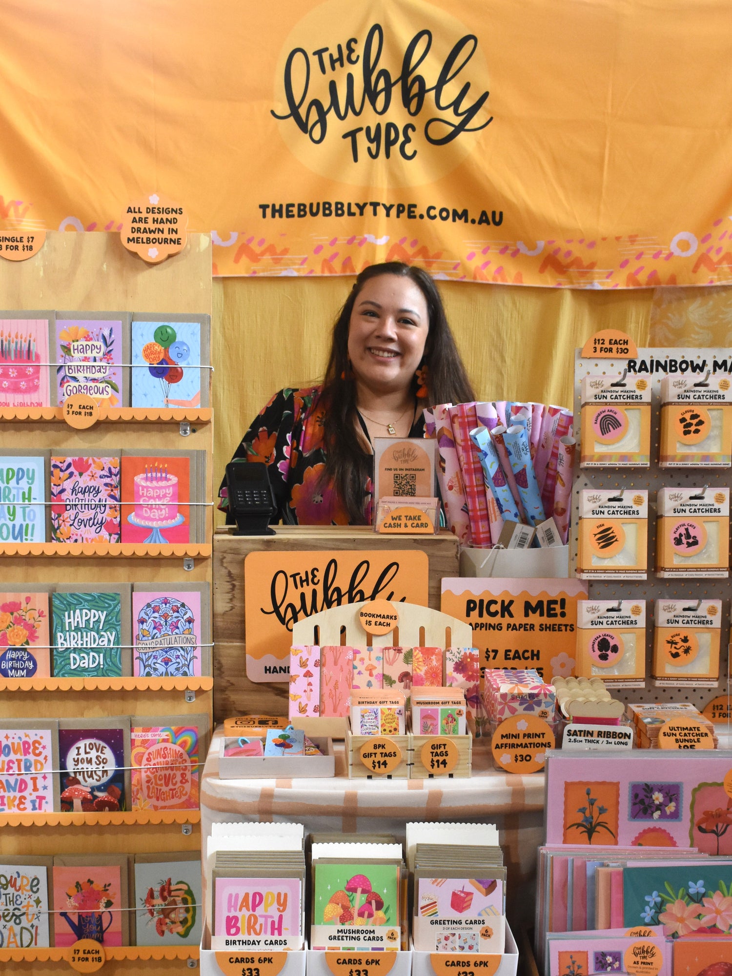 A smiling woman named Nicole stands behind a display of greeting cards and paper goods under a orange yellow canopy reading 'The Bubbly Type' and 'TheBubblyType.com.