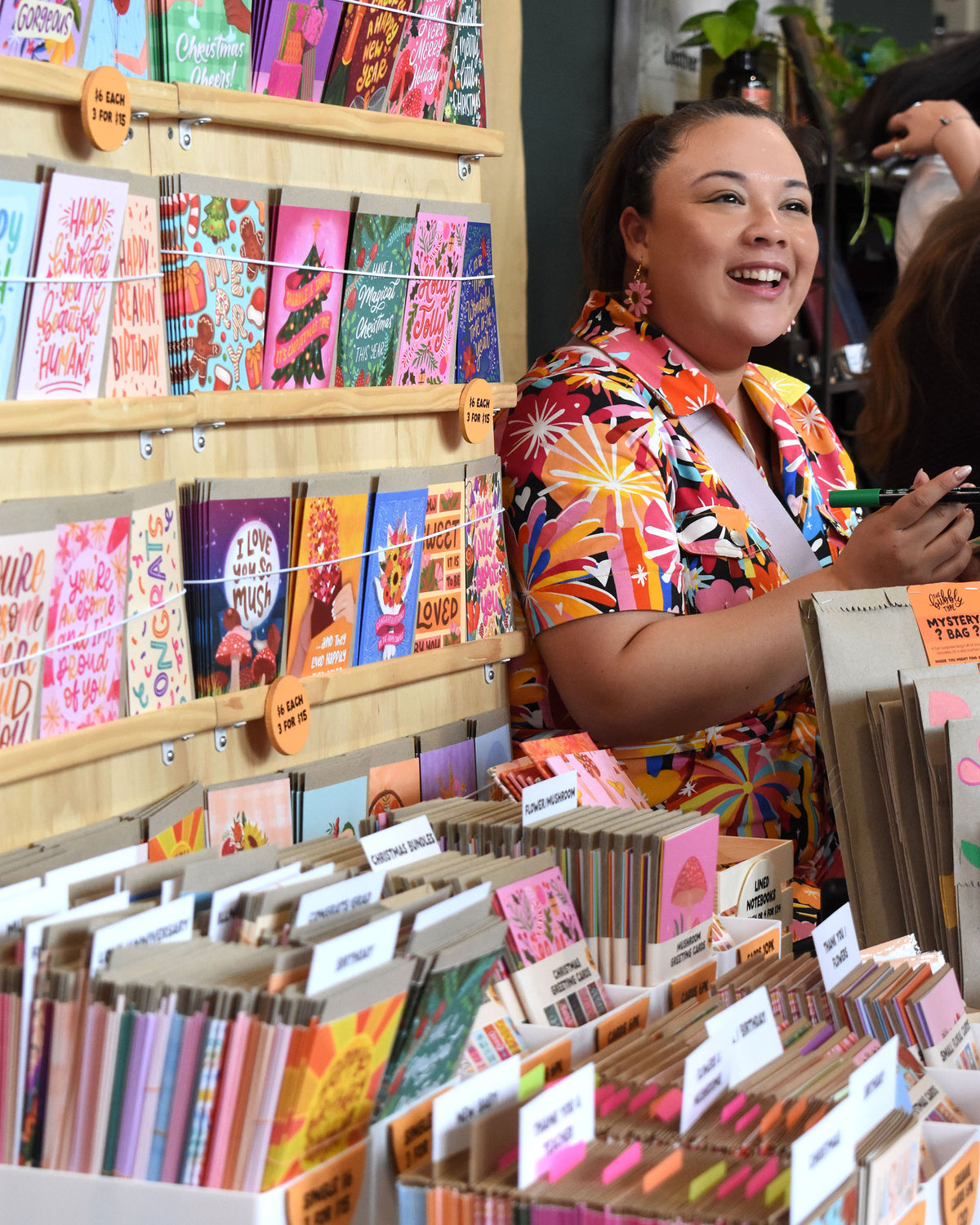 Nicole smiling in a colourful fireworks print jumpsuit stands behind a display of greeting cards and stationery in a shop setting.