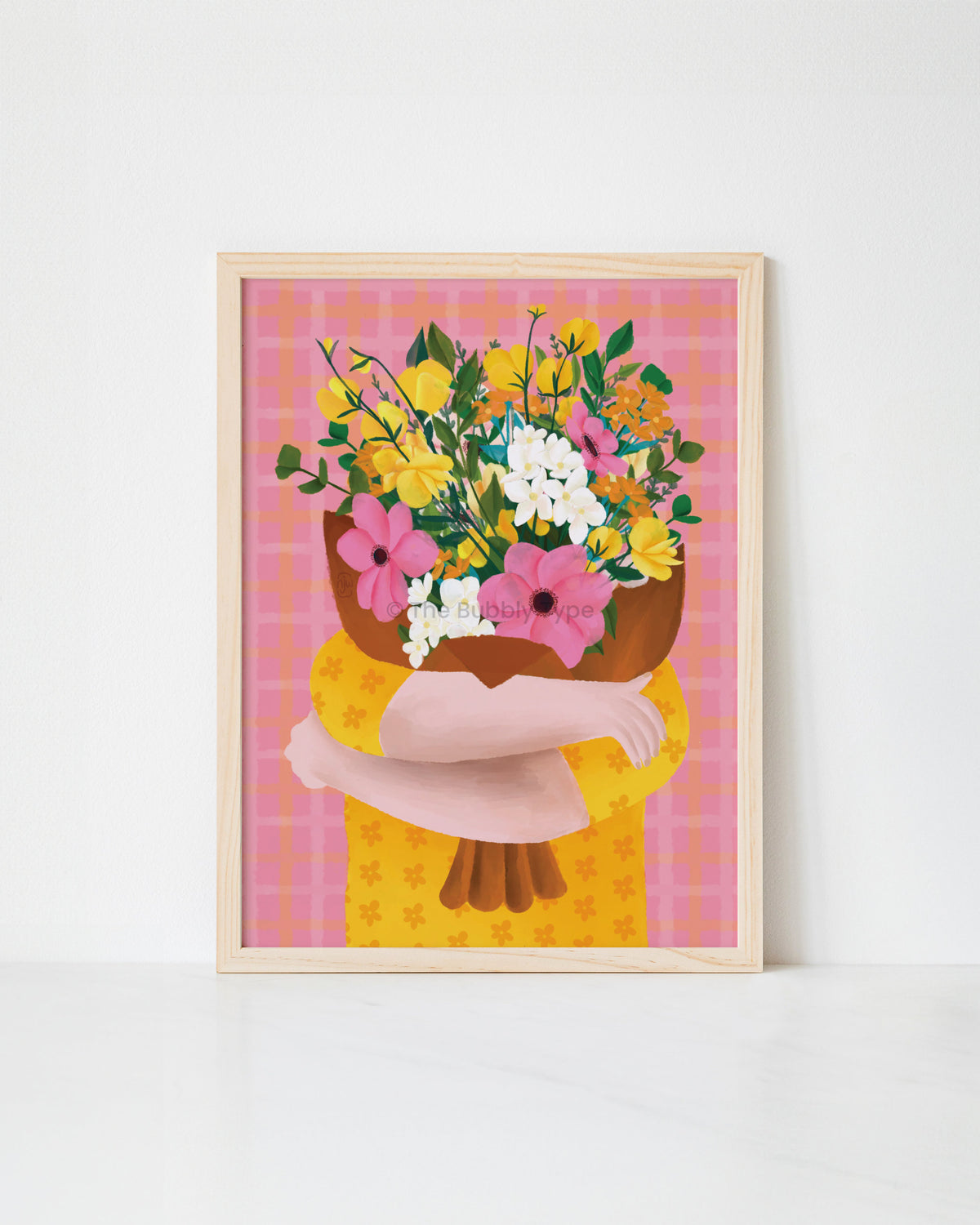 A framed art print featuring a stylized illustration of a person hugging a large bouquet of yellow, pink, and white flowers against a pink plaid background, shown leaning against a white wall.