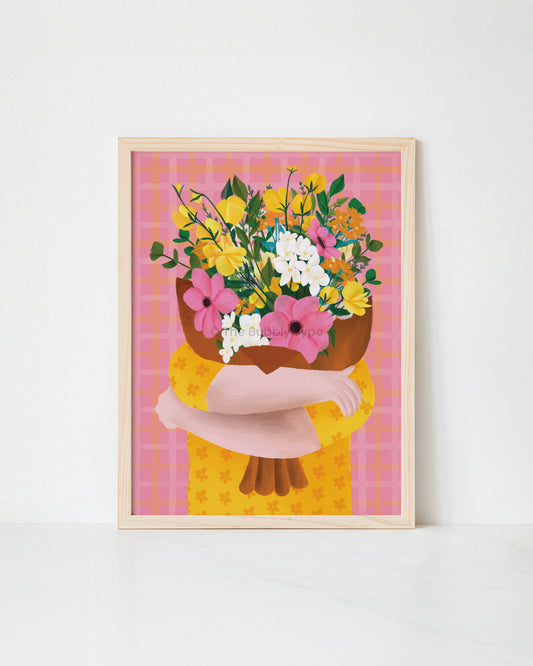 A framed art print featuring a stylized illustration of a person hugging a large bouquet of yellow, pink, and white flowers against a pink plaid background, shown leaning against a white wall.