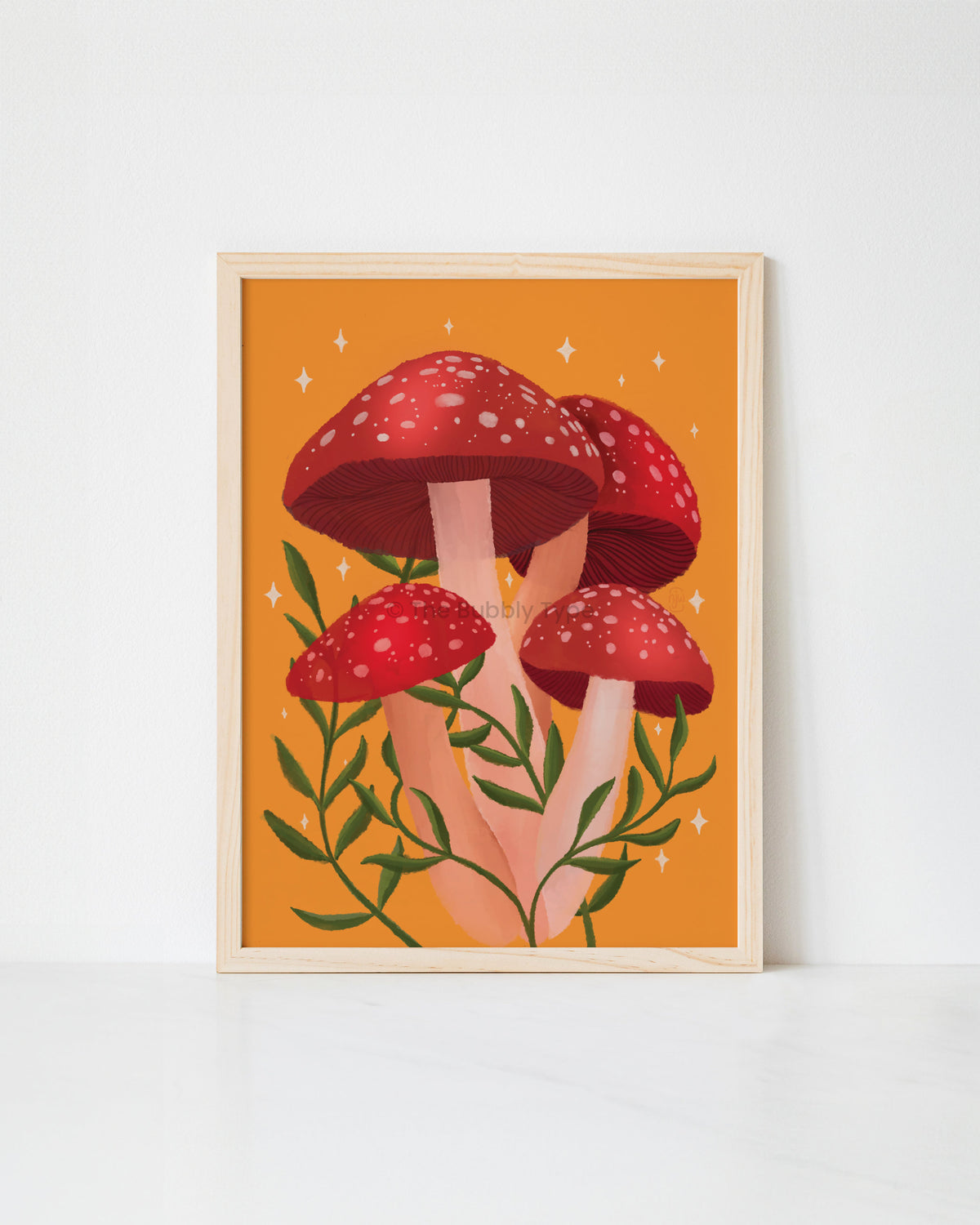 A framed art print featuring a whimsical illustration of four red mushrooms with white spots and green leaves on a warm orange background, displayed against a clean white wall.