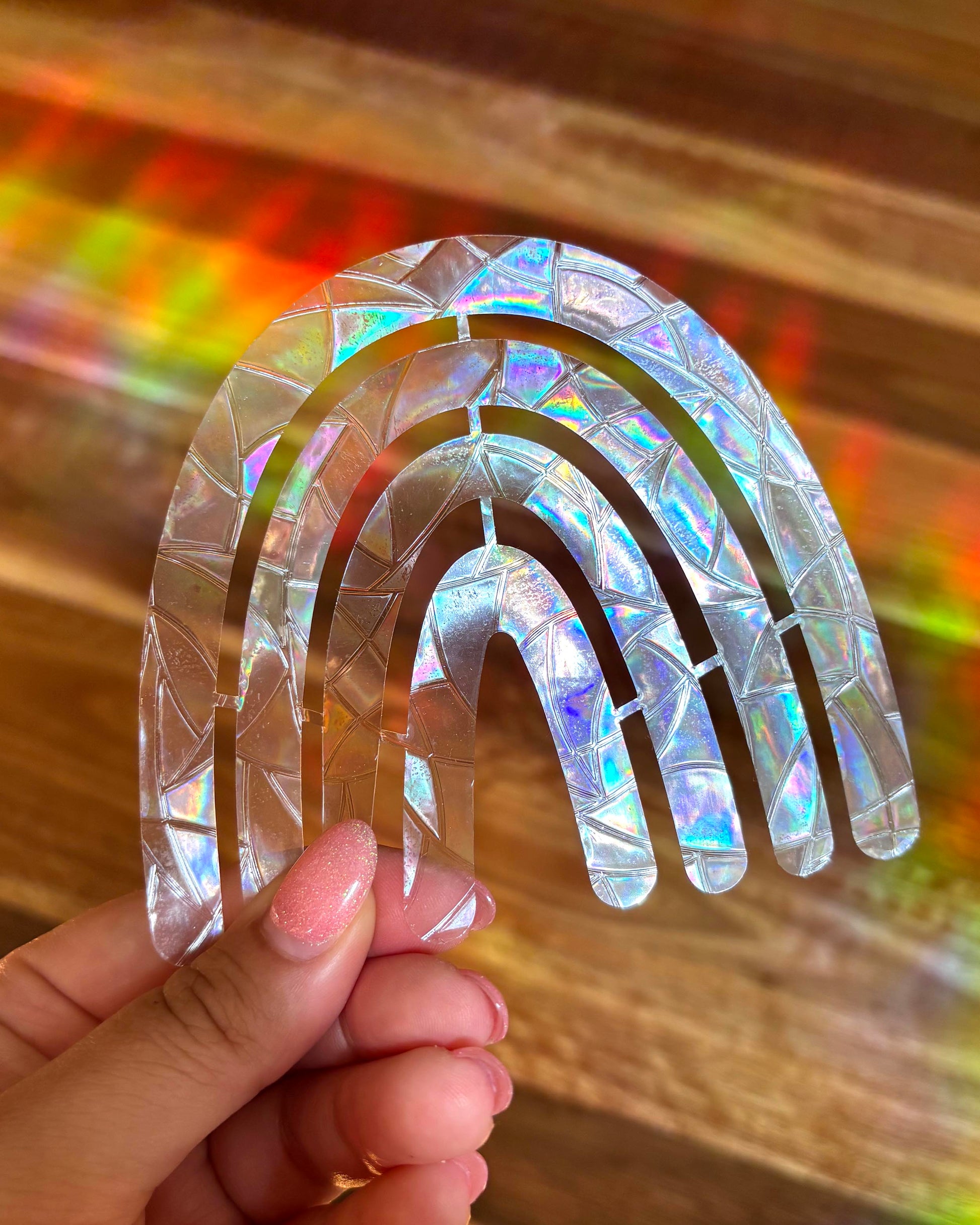 Holographic rainbow-shaped sun catcher window sticker held by a hand with a colourful background