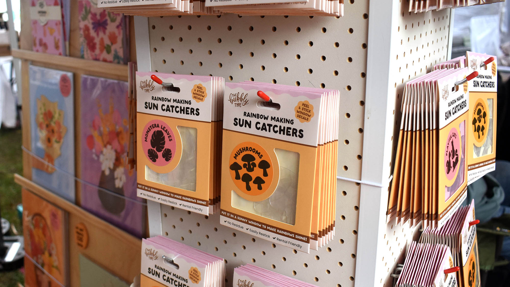 A close up of our sun catcher packs displayed at the market; specifically featuring our monstera leaf sun catchers and mushroom shaped sun catchers. some of our colourful art prints are visible out of focus to the left.