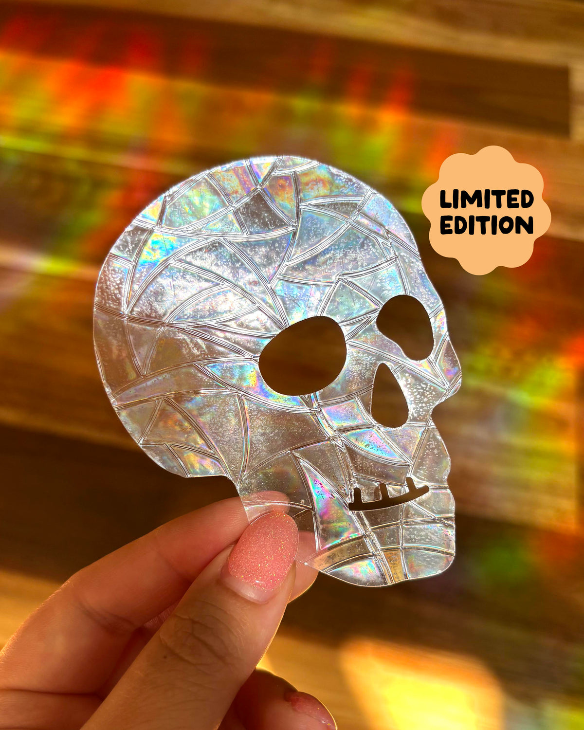 Holographic skull-shaped sun catcher window sticker held by a hand with a colourful rainbow lights, blurred background. A badge in the top right corner reads 'LIMITED EDITION'.