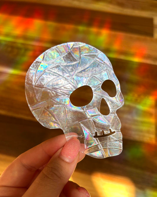 Holographic skull-shaped sun catcher held by a hand with a colourful rainbow lights, blurred background