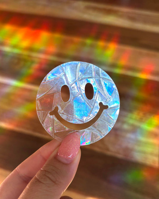 Holographic smiley face sun catcher window sticker held by a hand with a colourful blurred background