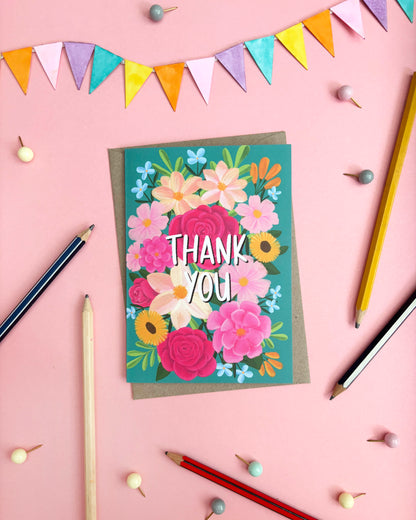 A colourful floral thank you card with the words 'THANK YOU' in white, layered over a brown envelope, styled on a pink background with coloured pencils, bunting, and small decorative beads.