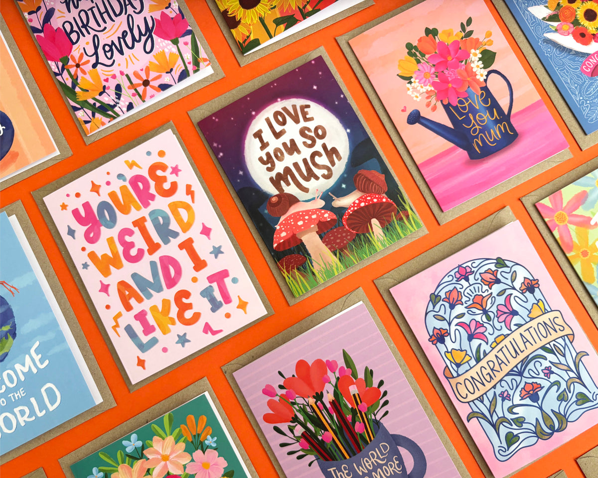 A close-up, angled view of a collection of colourful greeting cards arranged in a grid on an orange background. The cards feature various designs, including floral illustrations, mushrooms, a watering can, and playful text like 'You're Weird and I Like It' and 'I Love You So Mush'.