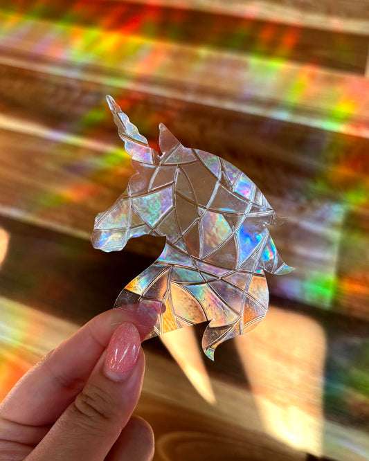 Iridescent unicorn-shaped sun catcher window cling sticker held by a hand with a colourful rainbow light burst  blurred in the background.