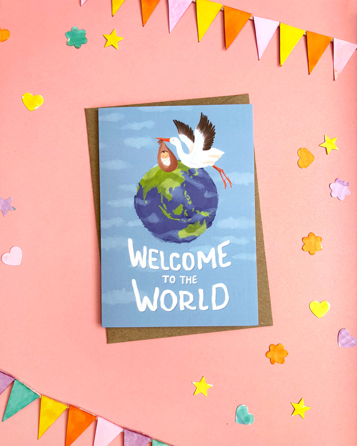 A greeting card with a blue sky and globe illustration, featuring a white bird carrying a baby and the text 'WELCOME TO THE WORLD' in white. The card is placed on a brown envelope against a pink background decorated with colourful paper flags and hearts.
