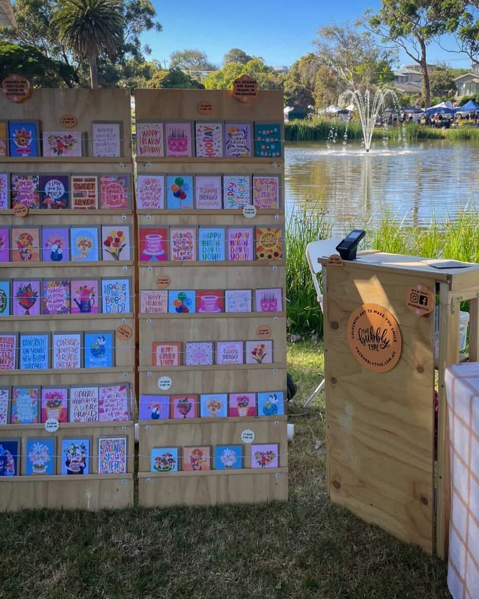 Outdoor market display with wooden boards covered in colorful handmade cards or stickers, a small wooden table with a round logo plaque, and a table covered in packaged items beside a grassy area and pond in the background.